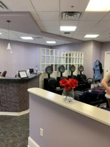 Salon room with four hair dryer chairs lined up against the back wall and a woman getting her hair washed in the foreground