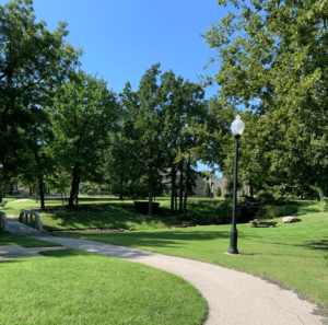 Curved outdoor walking path surrounded by green grass and trees with a small bridge over a ravine