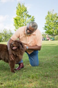 Man at dog park kneeling on the grass and petting brown dog