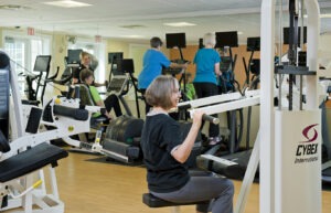 Woman using weight machine in fitness center with other people walking on elliptical machines in the background