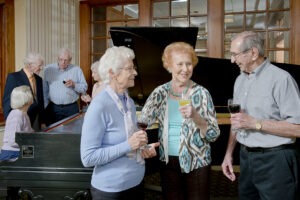 Group of men and women gathered around a piano holding wine glasses and talking to each other