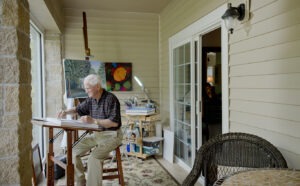 Man sitting and painting in covered porch studio with paintings hanging in the background