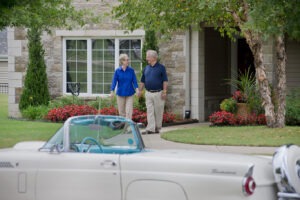 Man and woman holding hands and walking out of residence towards tan convertible car